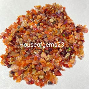 May include: A pile of small, irregularly shaped gemstones in shades of orange, brown, and red. The gemstones have a rough, textured appearance and reflect light, with the text "Houseofgems23" visible.