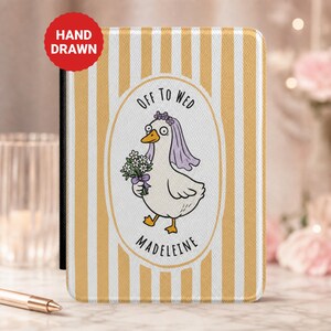 May include: A rectangular passport holder featuring a yellow and white vertical striped pattern. The center displays an oval frame containing a cartoon duck wearing a purple veil and holding a bouquet of flowers, with the text OFF TO WED MADELEINE.