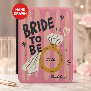 May include: A rectangular pink passport holder featuring vertical stripes and the text BRIDE TO BE in black. It displays a gold ring illustration with a diamond, a white veil, the year 2026, a small heart, and the name Madeleine in script.