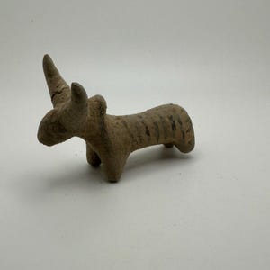 May include: A small, brown clay figurine of a bull with horns. The figurine is standing on four legs and has a striped pattern on its body.
