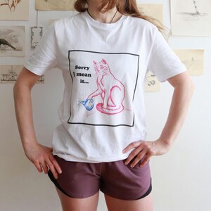 Może przedstawiać: Biała koszulka z różową ilustracją kota trzymającego niebieski przedmiot, oprawiona w czarny prostokąt. Tekst "Sorry I mean it..." znajduje się nad kotem. Osoba ma na sobie bordowe szorty z czarnym wykończeniem.
