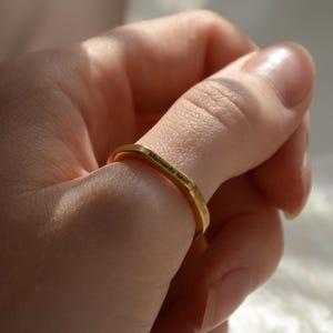 Peut inclure: Une bague dorée au design géométrique est portée au doigt. La bague a un dessus et des côtés plats. L'arrière-plan comprend une sculpture blanche et un éclairage doux.