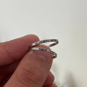 Puede incluir: Dos anillos de plata con las palabras "Memento Mori" y "Amor Fati" grabadas.