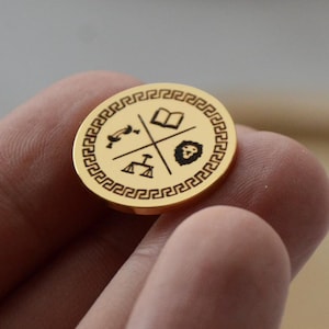 Peut inclure: Une pièce de monnaie dorée avec une bordure grecque et quatre symboles : un livre, un lion, une clé et une balance. La pièce est tenue entre les doigts, avec un plateau en bois flou en arrière-plan.