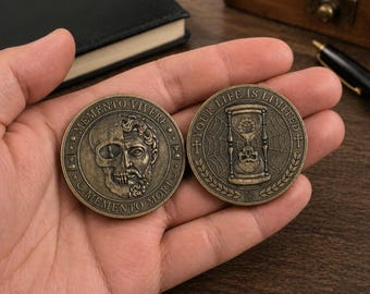 May include: Two antique bronze coins. One coin features a skull and a classical bust with the text "MEMENTO MORI" and "MEMENTO VIVERE". The other coin has an hourglass with the text "YOUR LIFE IS LIMITED".