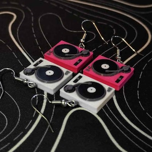 Puede incluir: Un par de pendientes con diseños de tocadiscos en miniatura. Un par es rosa con un disco negro, y el otro par es blanco con un disco negro.