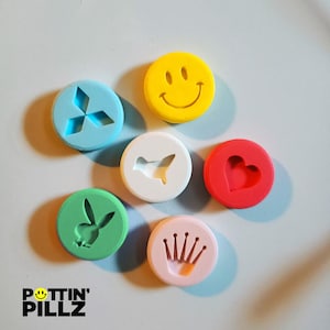 May include: Six colorful round plastic molds with different designs: a smiley face, a heart, a rabbit, a crown, a Mitsubishi logo, and a bird. The molds are on a light blue surface.