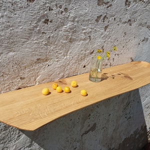 Peut inclure: Une étagère en bois avec un bord incurvé, montée sur un mur texturé rugueux. L'étagère est décorée d'un petit vase en verre rempli de fleurs jaunes et de plusieurs fruits jaunes.
