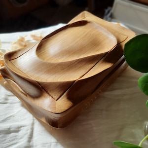 Op de afbeelding: Een houten doos met een uniek, golvend ontwerp. De doos is gemaakt van lichtbruin hout en heeft een gladde, gepolijste afwerking.