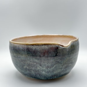 Könnte beinhalten: Eine handgefertigte Keramikschale mit einer einzigartigen, organischen Form. Die Schale hat eine Mischung aus blauen, grauen und violetten Farbtönen mit glänzendem Finish. Der Rand hat eine kontrastierende beige Farbe. Die Schale hat einen kleinen Ausguss an der Seite.