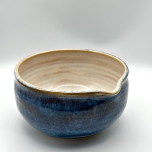 Könnte beinhalten: Handgefertigte Keramikschale mit blauem Außenbereich und hellbeigem Innenbereich. Die Schale hat einen kleinen Ausguss und eine glatte, glänzende Oberfläche. Geeignet zum Servieren von Speisen oder als dekoratives Element.