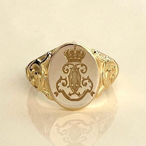 Könnte beinhalten: Ein goldener Siegelring mit ovaler Oberfläche, der ein detailliertes Wappen zeigt. Der Ring hat eine polierte Oberfläche und aufwendige Details am Band. Das Wappen enthält eine Krone und dekorative Schnörkel, was einen klassischen und eleganten Look erzeugt.