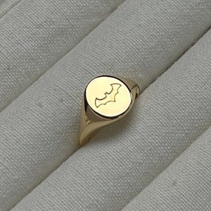 May include: A gold-colored signet ring with a bat emblem. The ring has a smooth, polished surface and a rounded face with the bat design. The ring is set against a white, textured background.