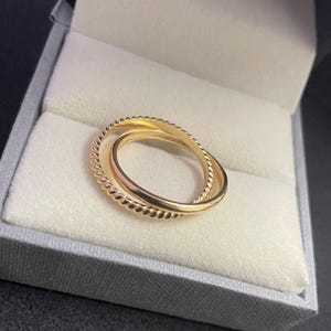 Könnte beinhalten: Ein goldfarbener Ring mit drei ineinandergreifenden Bändern, eines davon mit einer seilartigen Textur. Der Ring wird in einer grauen Schmuckschatulle mit weißem Innenraum präsentiert. Der Ring ist ein Schmuckstück.