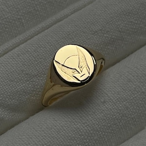 May include: A gold signet ring with a circular face featuring a stylized graphic. The ring is made of polished gold and sits on a white fabric surface. The graphic appears to be a superhero symbol.