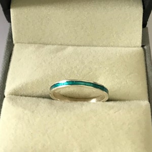 Puede incluir: Un fino anillo de banda de plata con una incrustación de esmalte verde turquesa. El anillo está en una caja de anillo de terciopelo blanco con las palabras "Allure Jewelcraft" y la dirección del sitio web "www.allurejewelcraft.com" impresas en el interior de la caja.