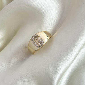 Puede incluir: Un anillo de sello dorado con la letra mayúscula "G" incrustada con pequeñas piedras preciosas transparentes. El anillo se presenta sobre una tela blanca y sedosa. El diseño del anillo es simple pero elegante. Una joya clásica.