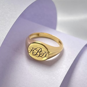 May include: A polished gold signet ring featuring a smooth, oval-shaped face. The centre of the ring is engraved with the letters HBD in a decorative, flowing script font. The band is solid gold with a rounded profile and a high-shine finish.