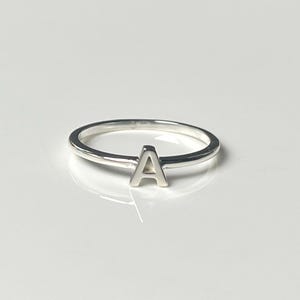 May include: A polished silver ring featuring a single capital letter A centred on the thin, circular band. The metal has a reflective, smooth finish, and the letter A is integrated directly into the top of the band, creating a simple, continuous design.