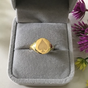 Pode incluir: Um anel de sinete de ouro com um design de paus. O anel está em uma caixa de anel de veludo cinza.