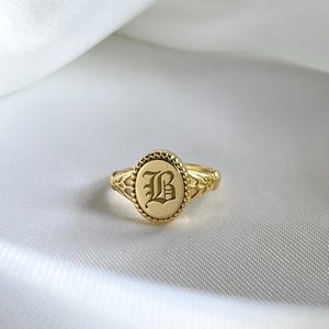 Op de afbeelding: Een goudkleurige zegelring met een ovaalvormig blad voorzien van een kralenrand en de letter B gegraveerd in een gotisch lettertype. De ringband is aan de zijkanten afgewerkt met gedetailleerde, bladachtige patronen in het metaal.