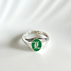 May include: A silver signet ring featuring an oval-shaped face with a vibrant green enamel inlay. Centered on the green background is a white letter L in a decorative gothic-style font. The band is polished silver with a smooth, rounded finish.
