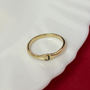 Könnte beinhalten: Ein goldfarbener Metallring mit einer glatten, polierten Ringschiene und einer flachen, kreisförmigen Oberfläche. Der Buchstabe J ist in einer einfachen, schwarzen, serifenlosen Schrift mittig auf der Ringfläche eingraviert.
