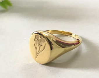 May include: A gold signet ring with an engraved floral design. The ring is oval shaped and has a smooth, polished finish.