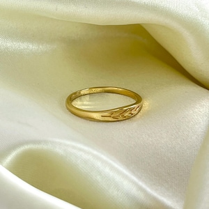 May include: A gold ring with a leaf design. The ring is set against a backdrop of a cream-coloured, silky fabric. The ring's band is thin and features an engraved leaf pattern on the front. The ring is a simple, elegant piece of jewellery.