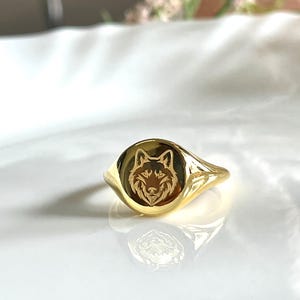 Könnte beinhalten: Ein polierter goldfarbener Siegelring mit einer runden Frontplatte, die ein detailliertes, eingraviertes Wolfskopfmotiv zeigt. Der Ring hat eine glatte, reflektierende Oberfläche und eine schmal zulaufende Ringschiene mit Spiegelung.