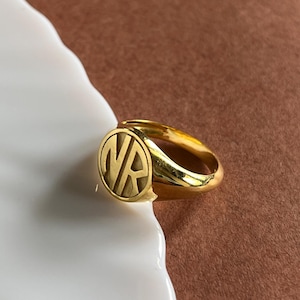 Può includere: Un anello a sigillo color oro con una parte superiore circolare che presenta le lettere NR in rilievo al centro. L'anello ha una superficie lucida e riflettente e una fascia liscia e arrotondata che si restringe verso la parte posteriore.
