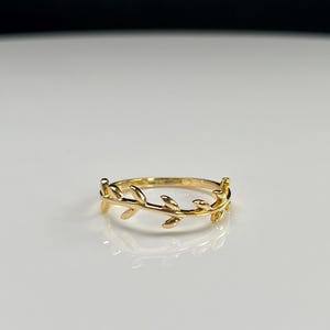 Op de afbeelding: Een gouden ring met een delicate band die is ontworpen om op een slanke wijnstok te lijken, met kleine, gepolijste blaadjes die zich langs de omtrek uitstrekken. Het metaal heeft een heldere, reflecterende afwerking die het licht opvangt.