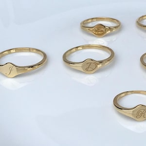 May include: Six gold signet rings with different initial letters engraved on the top. The rings are arranged in a semi-circle on a white surface.