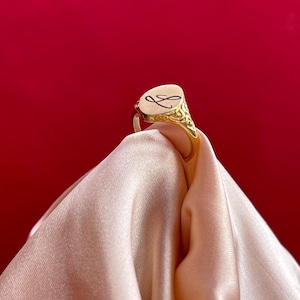 Puede incluir: Un anillo de sello dorado con una inicial grabada en una cara ovalada color crema. La banda del anillo presenta un diseño ornamentado y detallado. El anillo se muestra sobre un fondo rojo y una tela rosa claro drapeada.