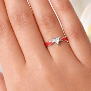 May include: A silver-toned ring featuring a central triangular setting with a small clear stone, flanked by vibrant red enamel inlay on both sides of the band. The ring is worn on a finger, showcasing the geometric design and the contrast between the metallic silver finish and the bright red color.