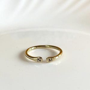 Puede incluir: Un anillo abierto de tono dorado con acabado pulido, que presenta la letra S grabada en un extremo y la letra M grabada en el otro. El anillo tiene un diseño circular delgado con un pequeño espacio entre los dos extremos, creando una apariencia simple y ajustable.
