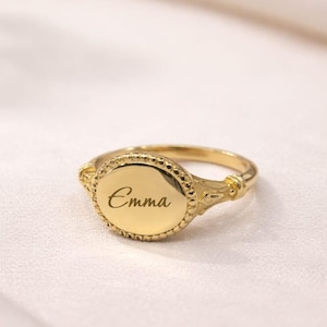 May include: A gold signet ring, featuring the name "Emma" inscribed on its oval face. The ring has an ornate design around the face and on the shoulders of the band. The ring is presented on a white, textured surface.