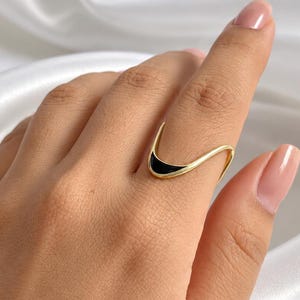 Puede incluir: Un anillo de tono dorado con un diseño de onda abierto y liso. El centro de la banda curva está acentuado con una incrustación negra elegante que sigue el contorno del metal, creando un contraste marcado con el acabado dorado pulido.