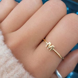May include: A thin gold ring featuring a single capital letter M as the centrepiece. The band is smooth and polished, showcasing a minimalist design that sits centred on the finger. The metal has a bright, reflective yellow gold finish.