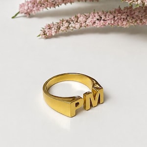 May include: A gold-toned metal ring featuring the capital letters PM prominently displayed on the front surface. The band has a smooth, polished finish and a solid construction, showcasing a clean design with no additional embellishments or stones.