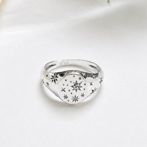 以下が含まれることがあります： 天体の星模様が刻印された楕円形のフェイスを持つシルバーのシグネットリング。リングのバンドとフェイスには、さまざまなサイズの星のモチーフが複数刻まれています。バンドの内側には925の刻印があります。
