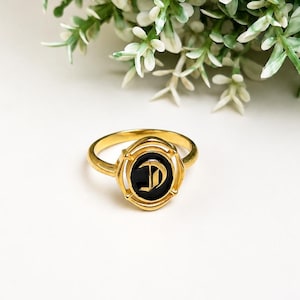 Pode incluir: Um anel de tom dourado com uma incrustação preta de formato oval ao centro. A incrustação preta exibe uma letra D estilizada em fonte gótica. O aro do anel é liso e circular, enquanto a cravação ao redor da incrustação tem uma moldura.