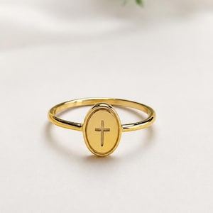 Könnte beinhalten: Ein goldfarbener Ring mit einer dünnen, polierten Ringschiene und einer zentralen, ovalen Platte. Die Platte ist mit einem vertikalen Kreuzsymbol graviert, das mittig im Oval platziert ist. Das Metall hat ein glattes, reflektierendes Finish.