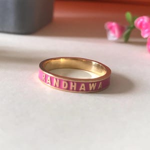 May include: A gold ring with a pink enamel band and the word "RANDHAWA" inscribed in gold lettering.