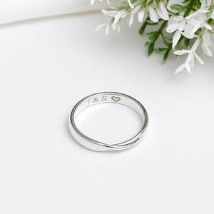 May include: A polished silver-toned band ring featuring a smooth, rounded surface. The interior of the ring is engraved with the text J & S followed by a small heart symbol. The ring has a simple, continuous circular design with a reflective finish.