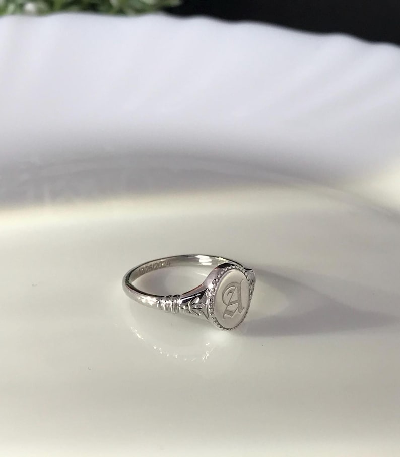 Pode incluir: Um anel de sinete de prata com uma parte superior ovalada que apresenta uma letra "A" gravada.