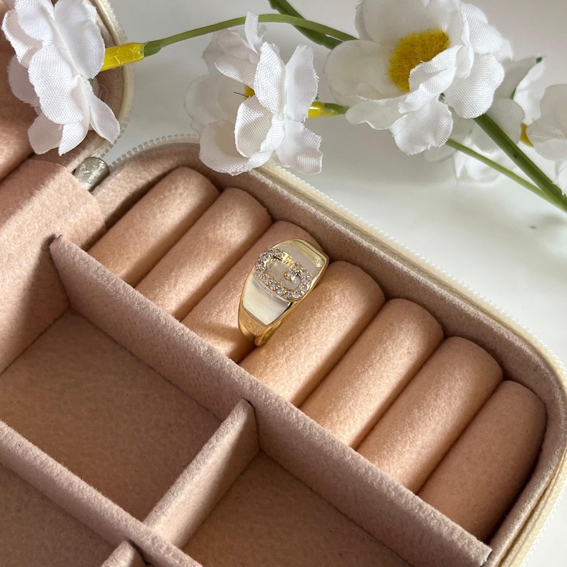 May include: A gold-colored ring with a white enamel face and a diamond-studded initial. The ring is displayed in a pink jewelry box, with white flowers in the background. The ring is a fashion accessory.