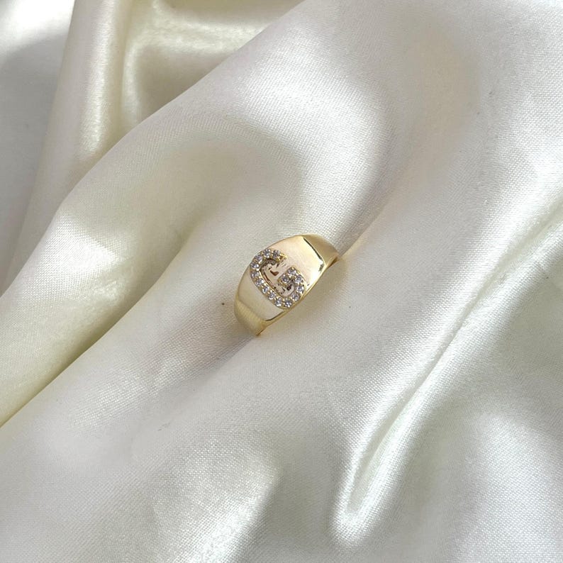 May include: A gold-colored signet ring with a capital letter "G" encrusted with small, clear gemstones. The ring is set against a backdrop of white, silky fabric, creating a luxurious feel. The ring's design is simple yet elegant.