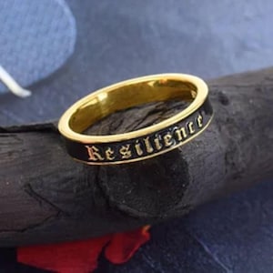 Puede incluir: Un anillo de banda de oro con un acabado de esmalte negro. El anillo tiene la palabra "Resilience" inscrita en letras doradas.
