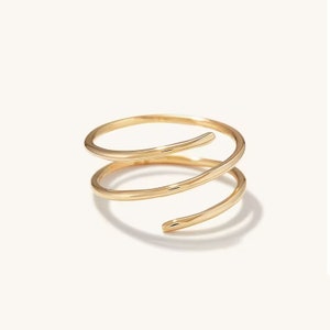 May include: A gold-colored spiral ring with three loops. The ring has a smooth, polished finish and is set against a white background. The design is minimalist and modern, suitable for everyday wear.
